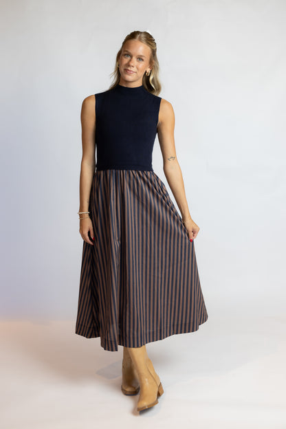 A woman modeling a sleeveless mock-neck midi dress with a dark navy top and a brown and navy vertically striped skirt, paired with tan ankle boots, against a light gray studio background.