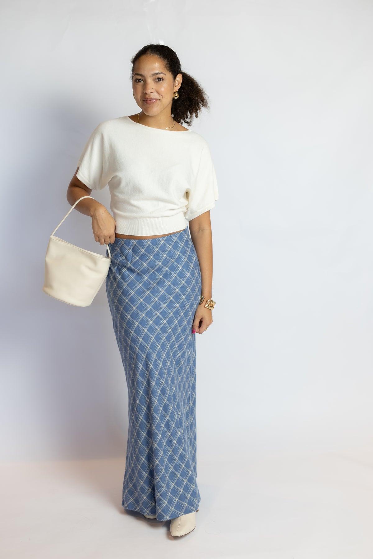 A woman modeling a white short-sleeve knit top and a blue plaid maxi skirt, holding a cream-colored bucket bag against a plain light gray studio background.