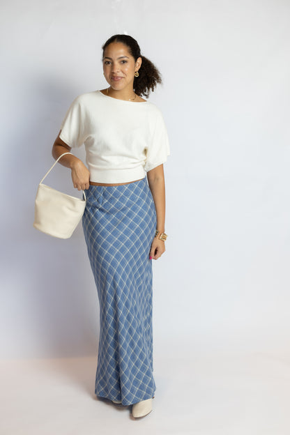 A woman modeling a white short-sleeve knit top and a blue plaid maxi skirt, holding a cream-colored bucket bag against a plain light gray studio background.
