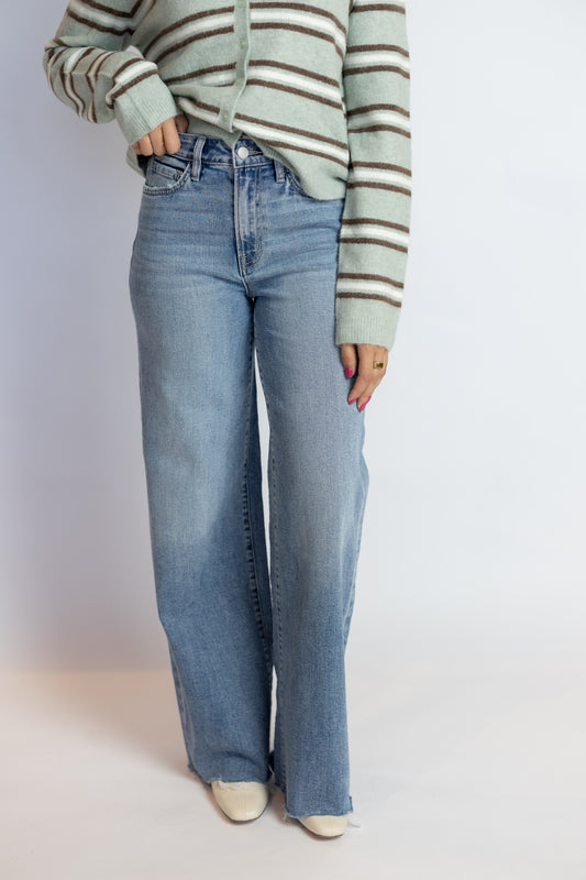 A person wearing light-wash blue wide-leg jeans, a sage green and brown striped cardigan, and cream pointed-toe heels, standing against a plain light gray studio background.