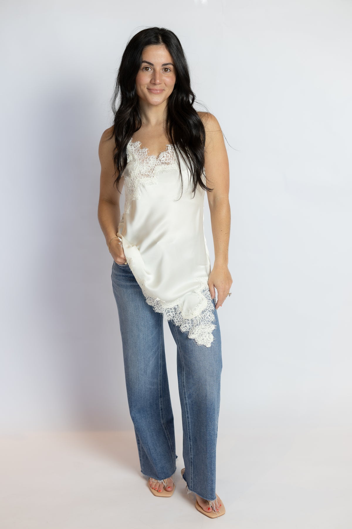 A woman modeling a white satin camisole top with lace trim, paired with blue jeans and tan sandals, standing against a plain white studio background.