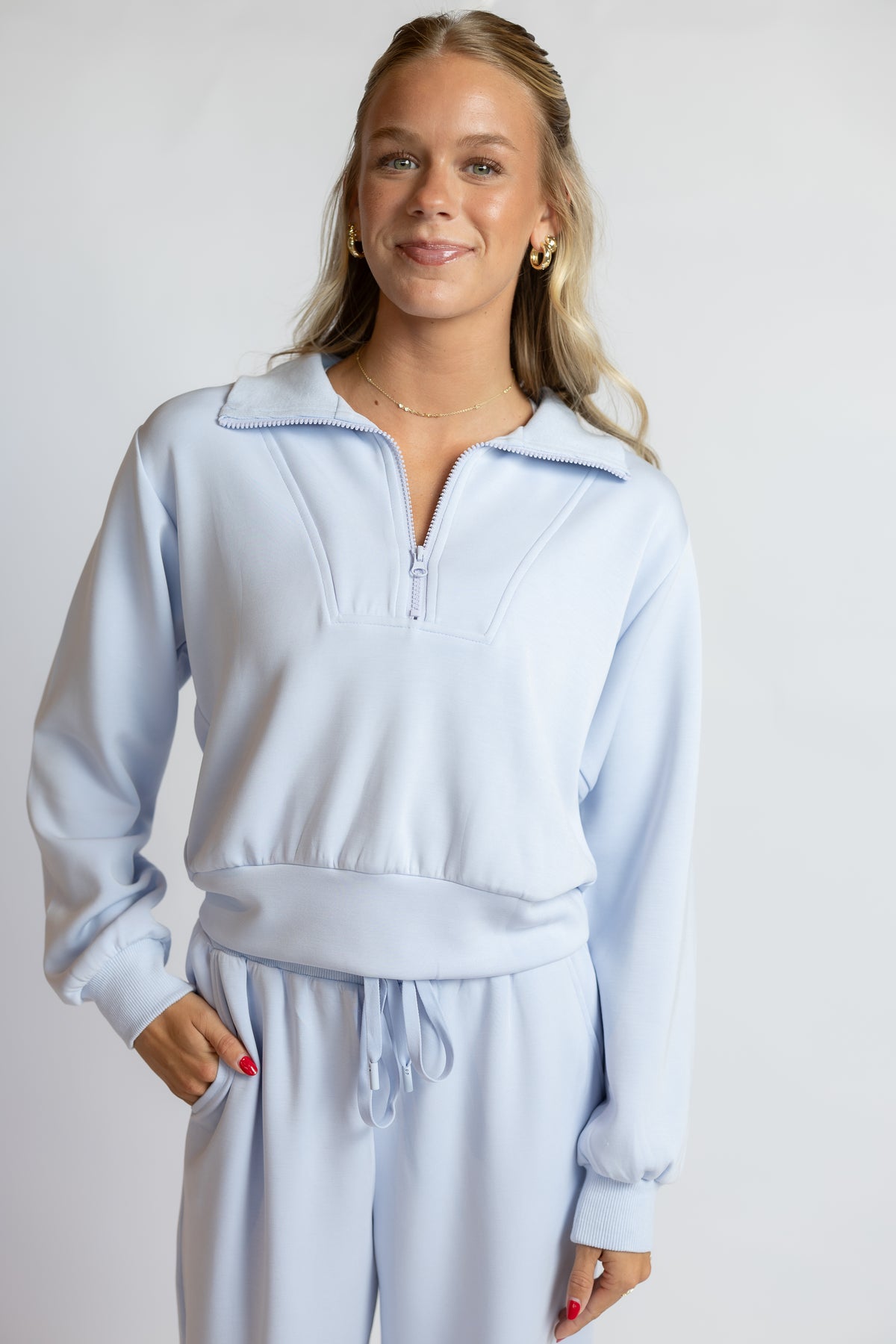 A woman modeling a light blue matching lounge set consisting of a long-sleeve half-zip sweatshirt and matching drawstring sweatpants, shot against a plain light gray studio background.