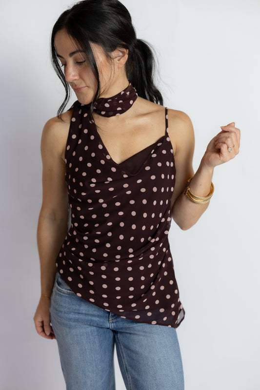 A woman wearing a dark brown halter top with beige polka dots and a matching neck scarf, paired with blue jeans, against a plain light gray background.