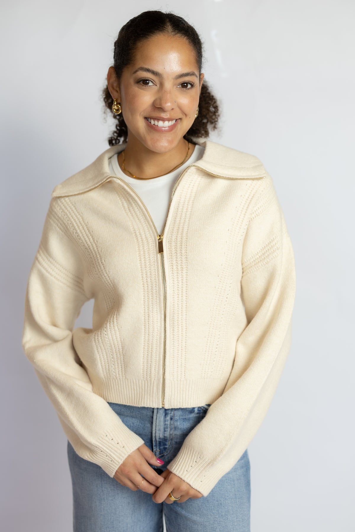 A woman wearing a cream-colored ribbed knit zip-up jacket with a wide collar, styled over a white t-shirt and blue jeans, standing against a light gray studio background.