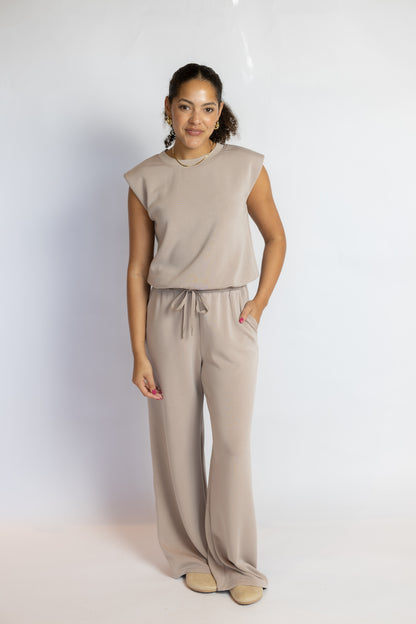 A woman modeling a matching two-piece lounge set consisting of a sleeveless top and wide-leg drawstring pants in a light taupe-beige color, set against a plain white studio background.