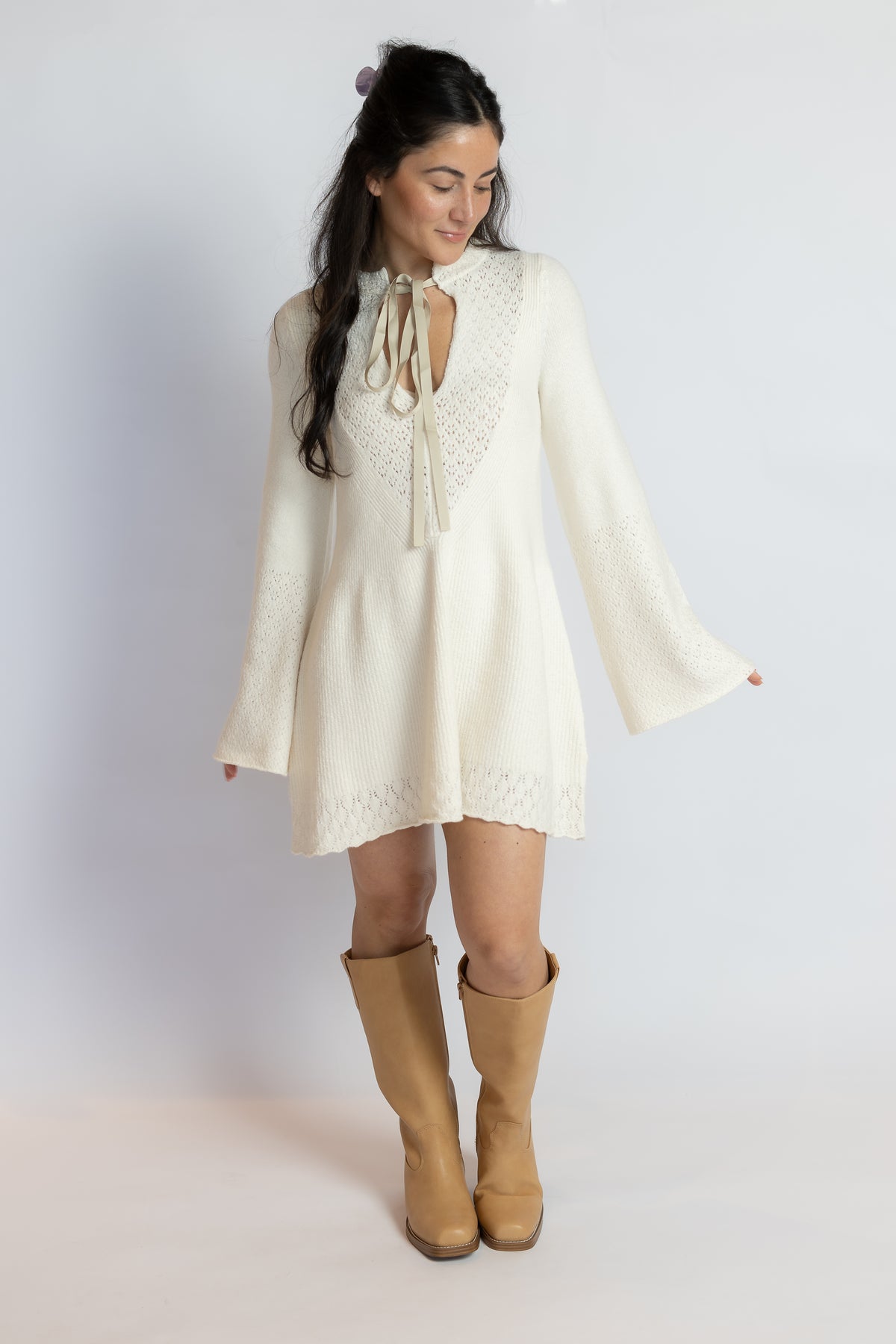 A woman modeling a cream-colored long-sleeve mini dress with a lace keyhole tie neckline, paired with tan knee-high boots, against a light gray studio background.
