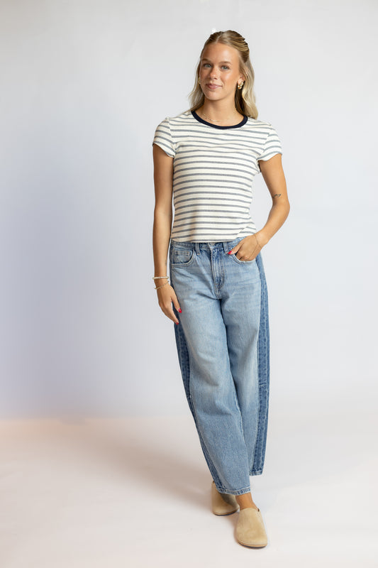 A woman modeling a short-sleeve striped t-shirt with dark navy trim and light blue wide-leg jeans with dark blue side panels, paired with beige slip-on shoes, against a light gray studio background.