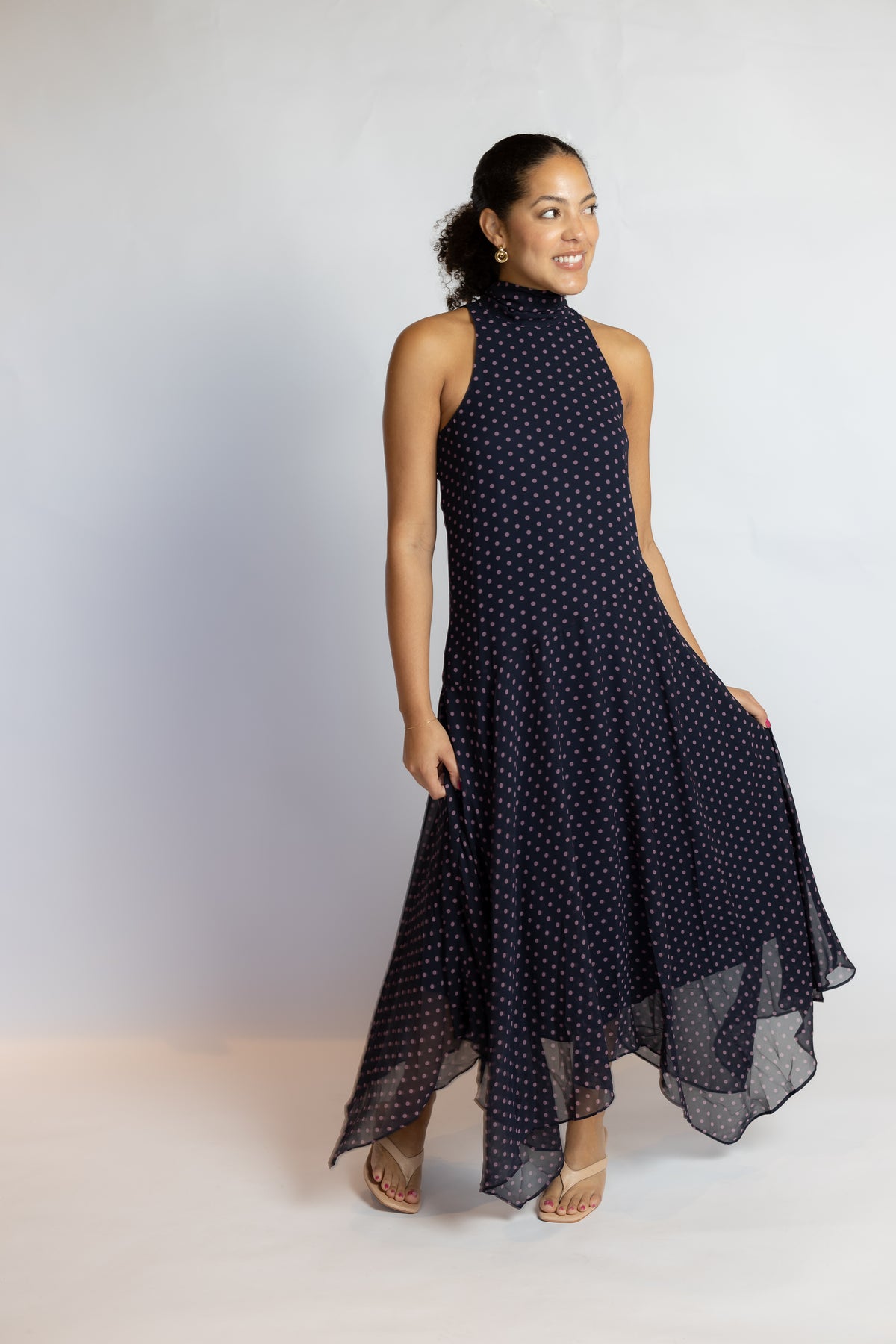 A woman modeling a sleeveless navy blue high-low dress with a white polka dot pattern, standing against a light gray studio background.