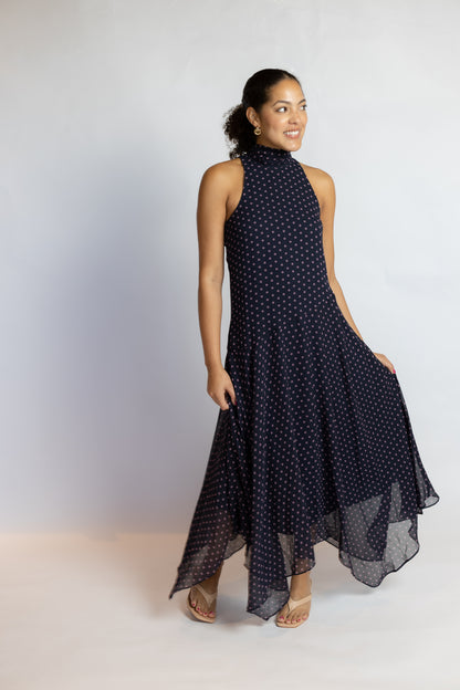 A woman modeling a sleeveless navy blue high-low dress with a white polka dot pattern, standing against a light gray studio background.