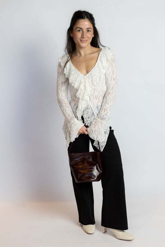 A woman modeling a white ruffled lace long-sleeve blouse paired with black wide-leg pants, holding a dark brown glossy handbag, against a light gray studio background.