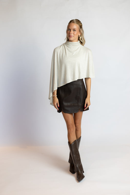 A woman modeling a cream-colored draped turtleneck top, a black leather mini skirt, and dark brown leather cowboy boots, standing against a plain light gray studio background.