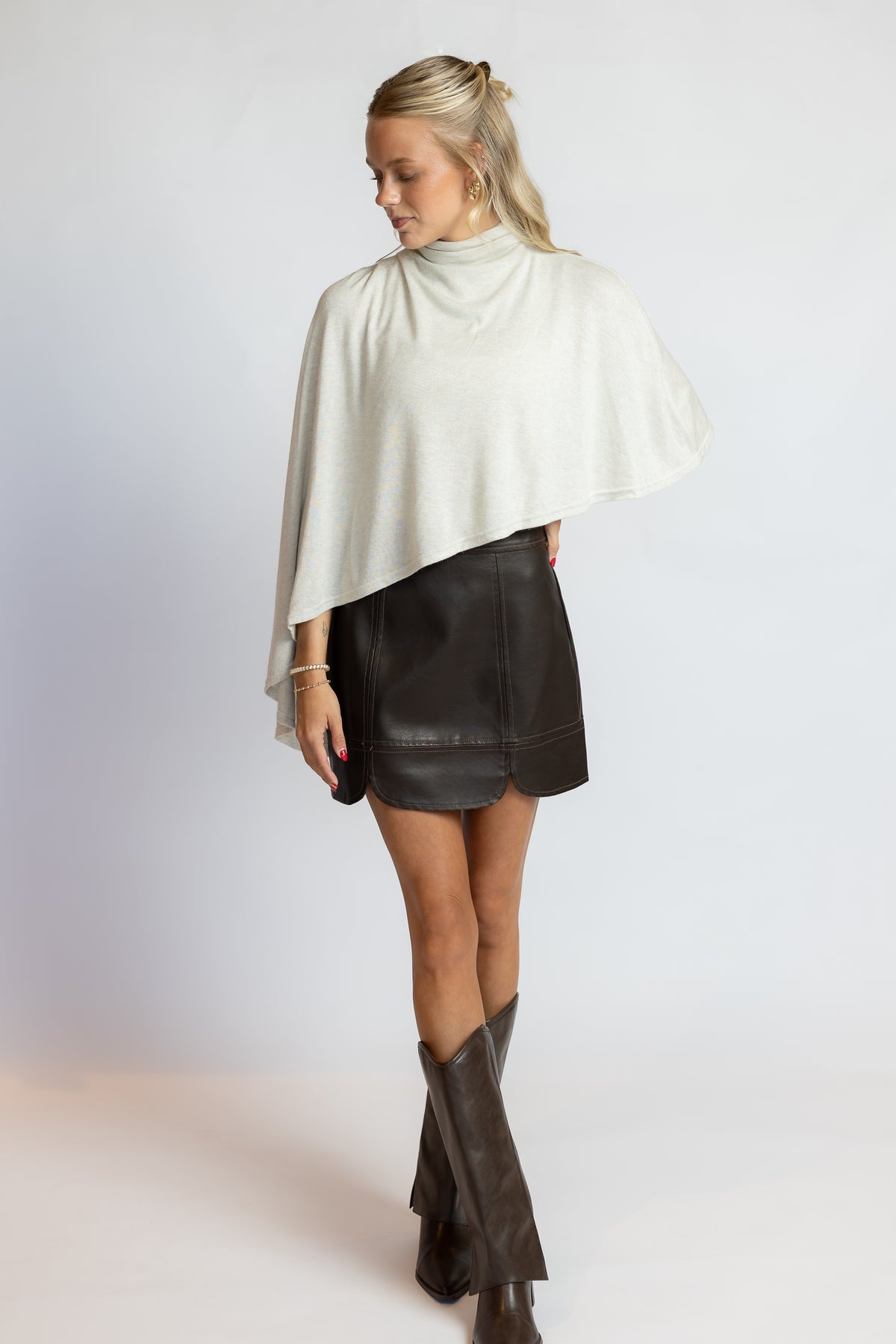 A woman modeling a cream-colored draped mock-neck cape top paired with a black faux-leather mini skirt and dark brown knee-high boots, photographed against a light gray studio background.