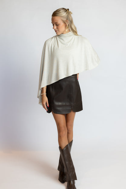 A woman modeling a cream-colored draped mock-neck cape top paired with a black faux-leather mini skirt and dark brown knee-high boots, photographed against a light gray studio background.