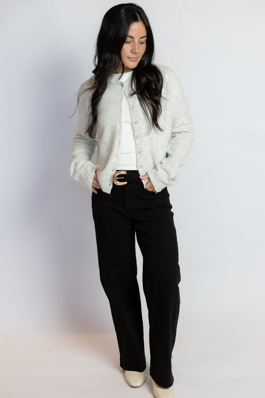 A woman modeling a light gray knit cropped cardigan over a white top, paired with black pants and beige heels, against a plain light gray studio background.