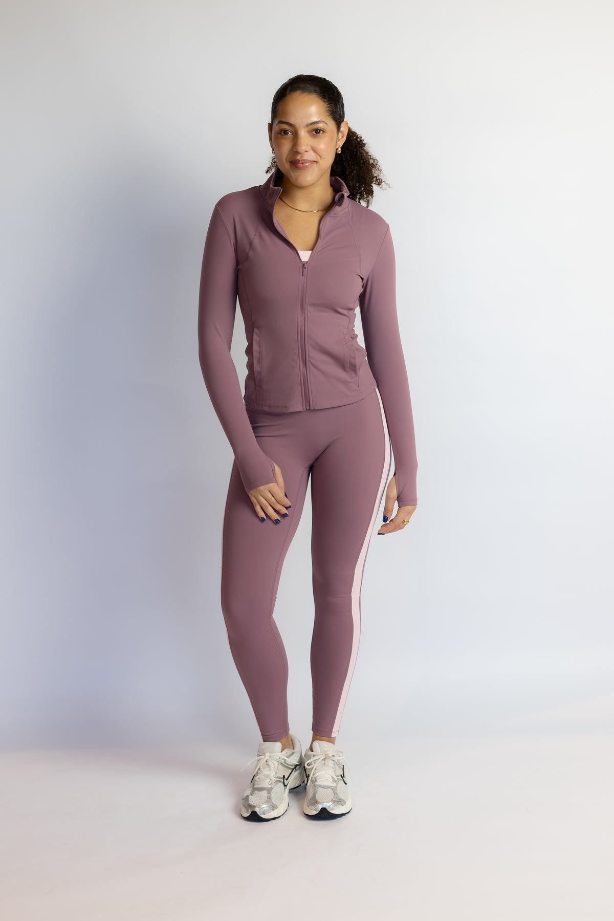 A woman modeling a matching athletic set consisting of a long-sleeve zip-up jacket and high-waisted leggings in a dusty mauve color, paired with white sneakers, against a light gray studio background.