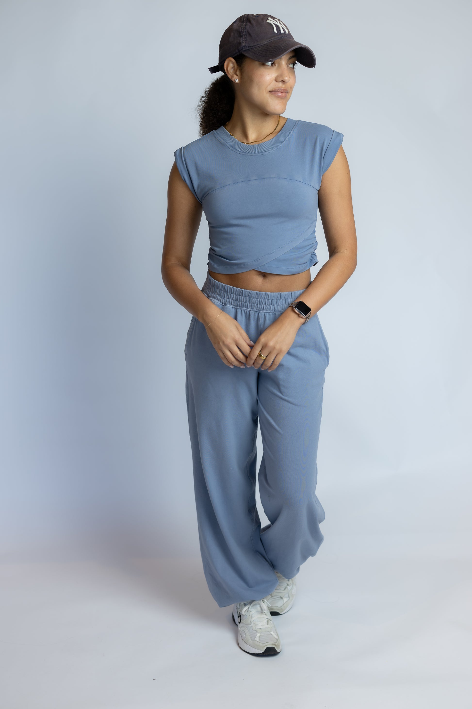 A woman modeling a matching dusty blue two-piece lounge set consisting of a cropped sleeveless top and wide-leg sweatpants, paired with a dark baseball cap and white sneakers, against a light gray studio background.