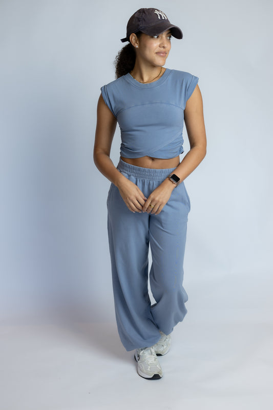 A woman modeling a matching dusty blue two-piece lounge set consisting of a cropped sleeveless top and wide-leg sweatpants, paired with a dark baseball cap and white sneakers, against a light gray studio background.