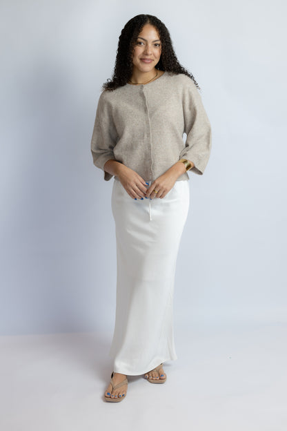 A woman modeling a light beige knit cropped sweater with three-quarter sleeves and a long white skirt, standing against a light gray studio background.