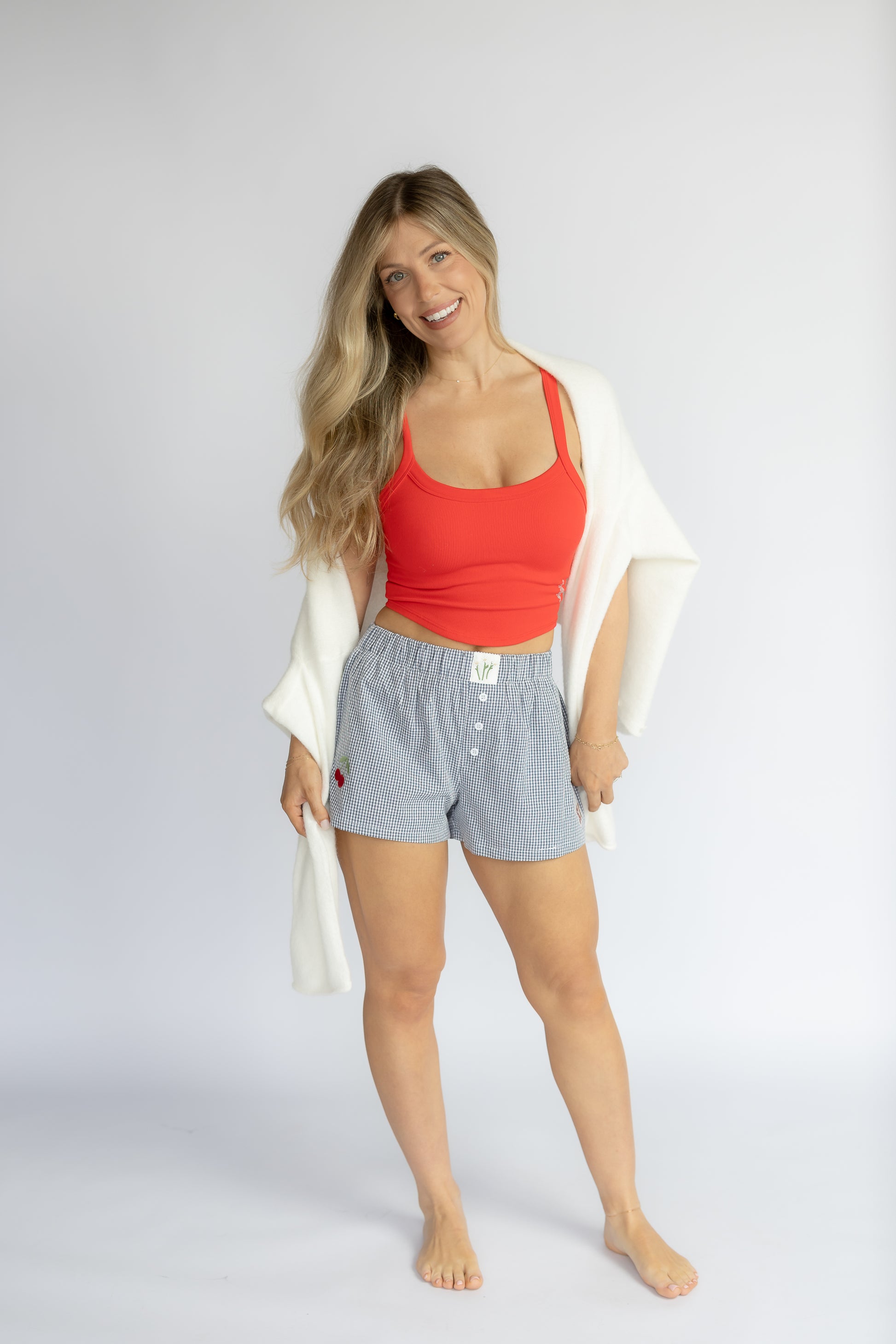 Woman wearing a red top and light blue shorts with a white cardigan on a plain background