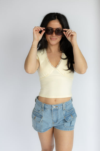 Woman wearing a yellow top and denim shorts, holding sunglasses against a white background