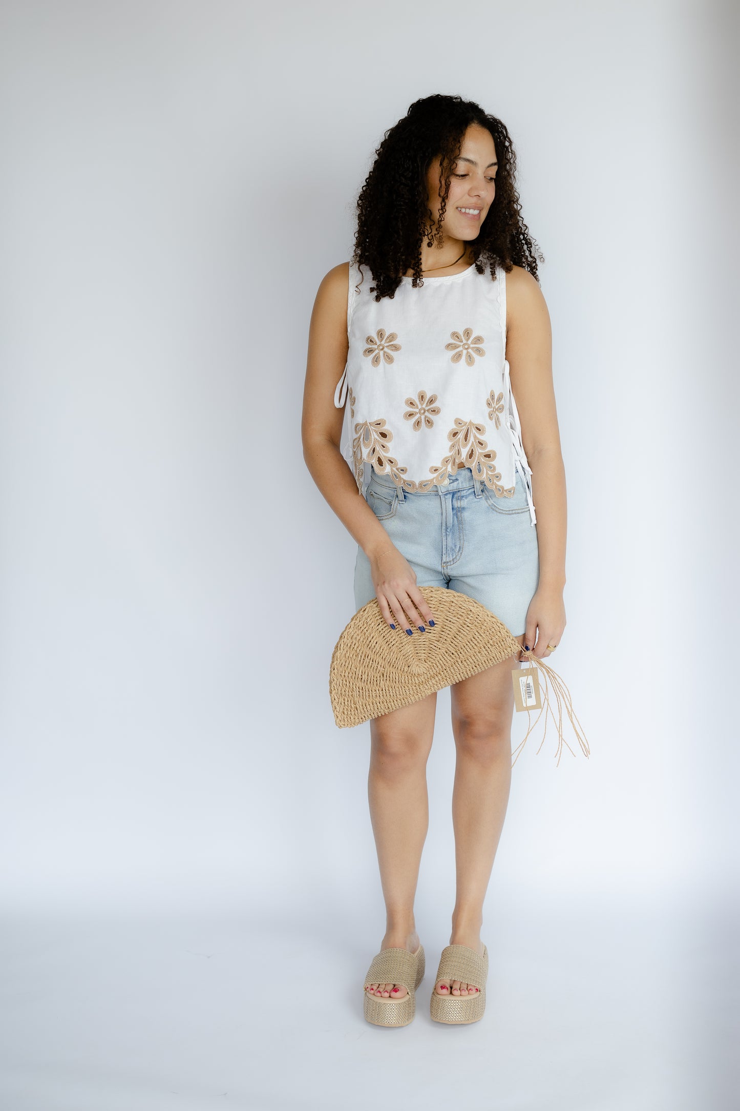 Woman wearing a sleeveless floral top and denim skirt with a straw clutch against a white background