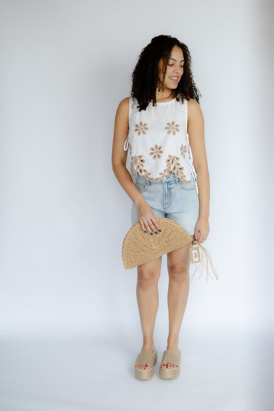 Woman wearing a sleeveless floral top and denim skirt with a straw clutch against a white background