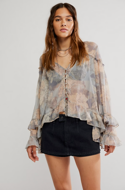 Woman wearing sheer long sleeve blouse with muted floral print, ruffle hem and sleeves, styled with black mini skirt