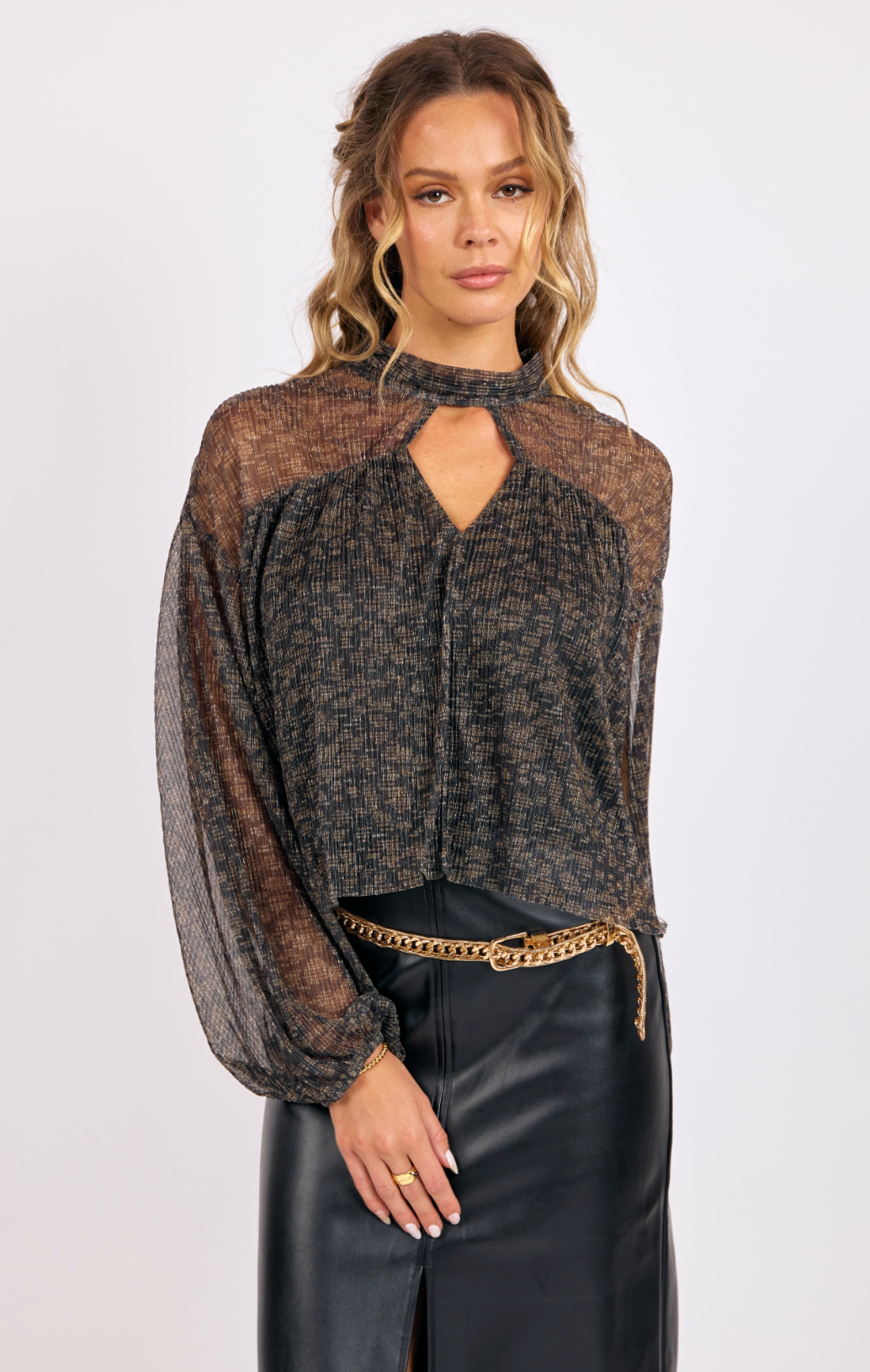 Woman wearing a metallic lurex keyhole blouse with sheer sleeves and a pleated texture paired with a black leather skirt and gold belt.