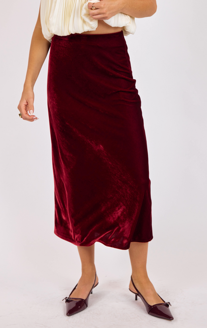 Woman wearing a deep red stretch velvet bias-cut midi skirt paired with an ivory pleated blouse and burgundy heels.