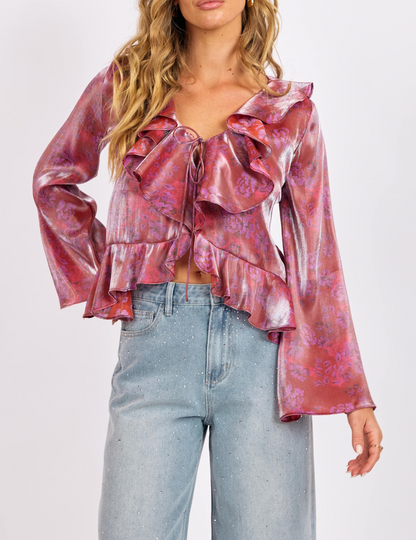 Woman wearing a shiny pink-lavender floral ruffle front top with tie-front closure and long sleeves, paired with light denim jeans.