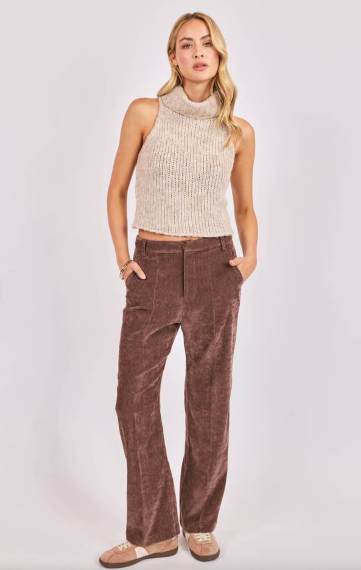 Woman wearing a sleeveless beige chunky knit turtleneck sweater with brown corduroy pants.
