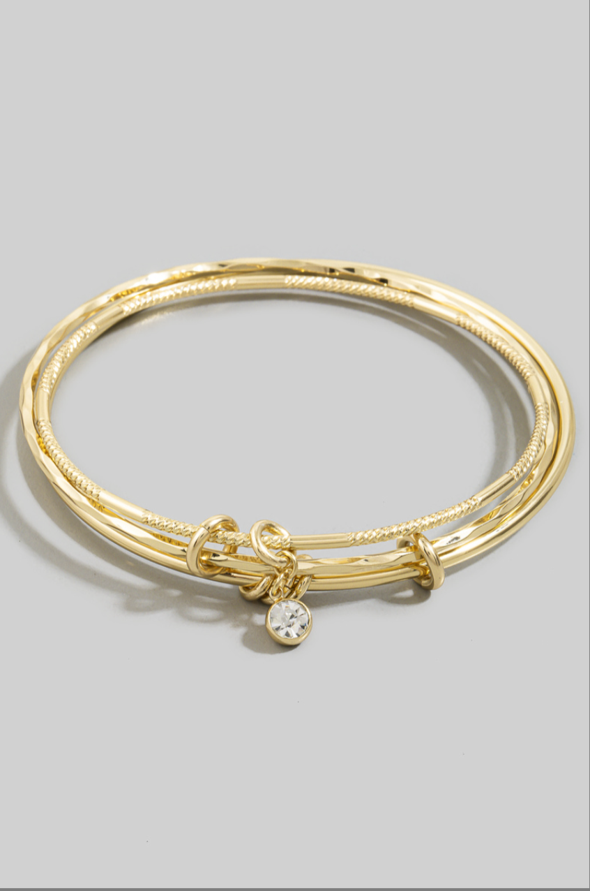 Three gold-tone textured bangles with knot accents and a dangling bezel-set cubic zirconia charm worn on a model's wrist.