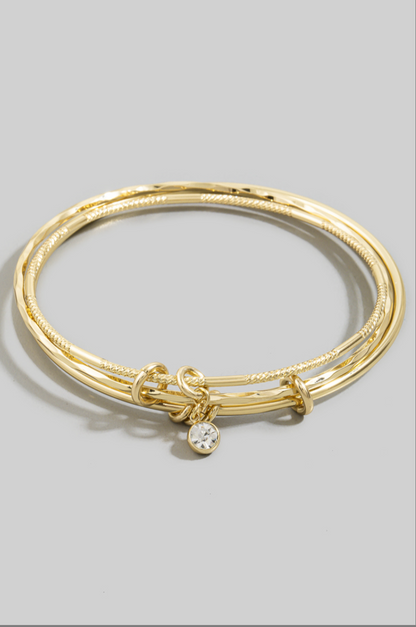 Three gold-tone textured bangles with knot accents and a dangling bezel-set cubic zirconia charm worn on a model's wrist.