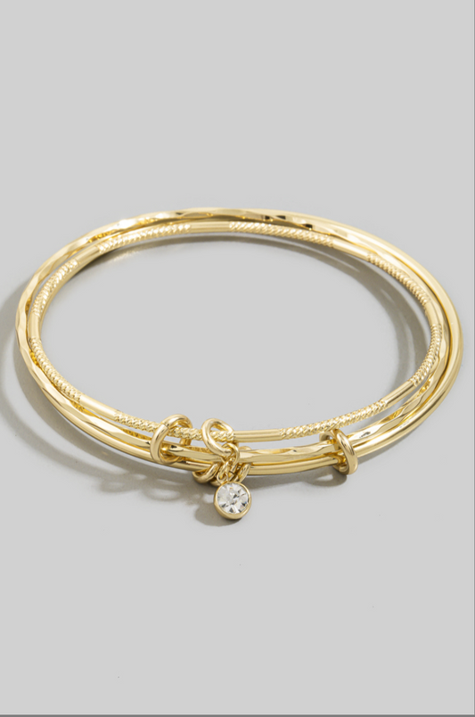 Three gold-tone textured bangles with knot accents and a dangling bezel-set cubic zirconia charm worn on a model's wrist.