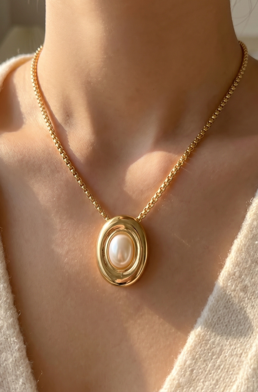Gold necklace featuring an oval pendant with a centered acrylic pearl on a polished chain.