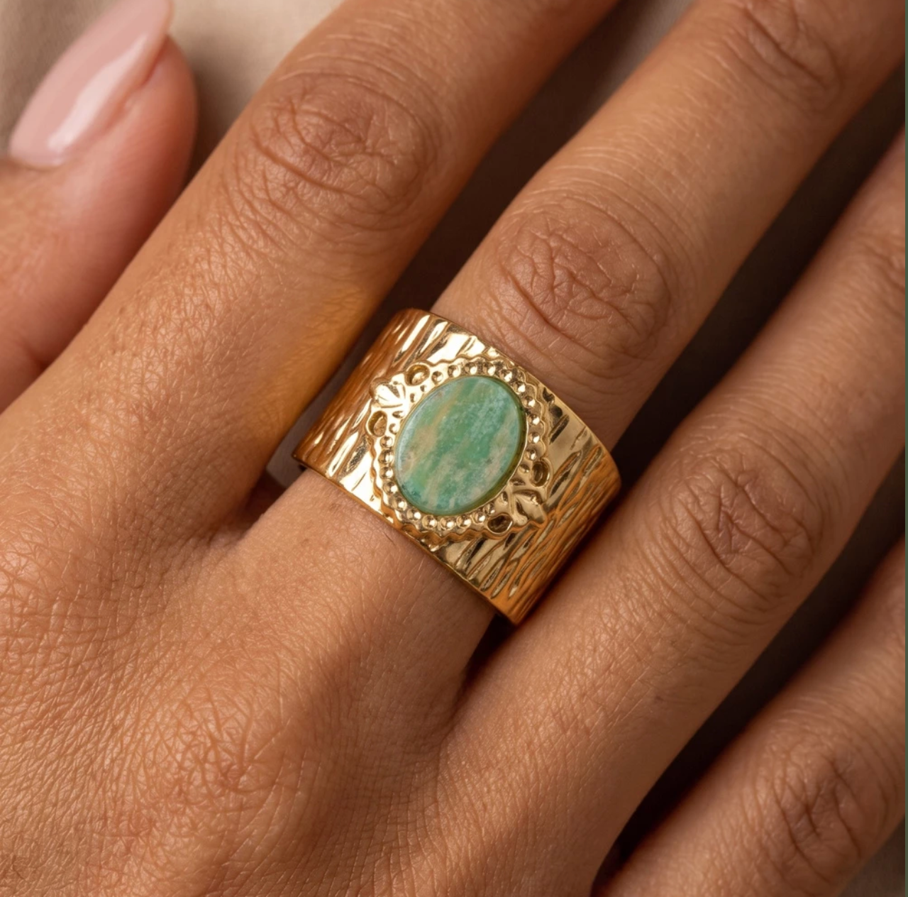Gold-tone stainless steel ring with a textured band featuring an oval green cabochon stone surrounded by a beaded halo and leaf accents.