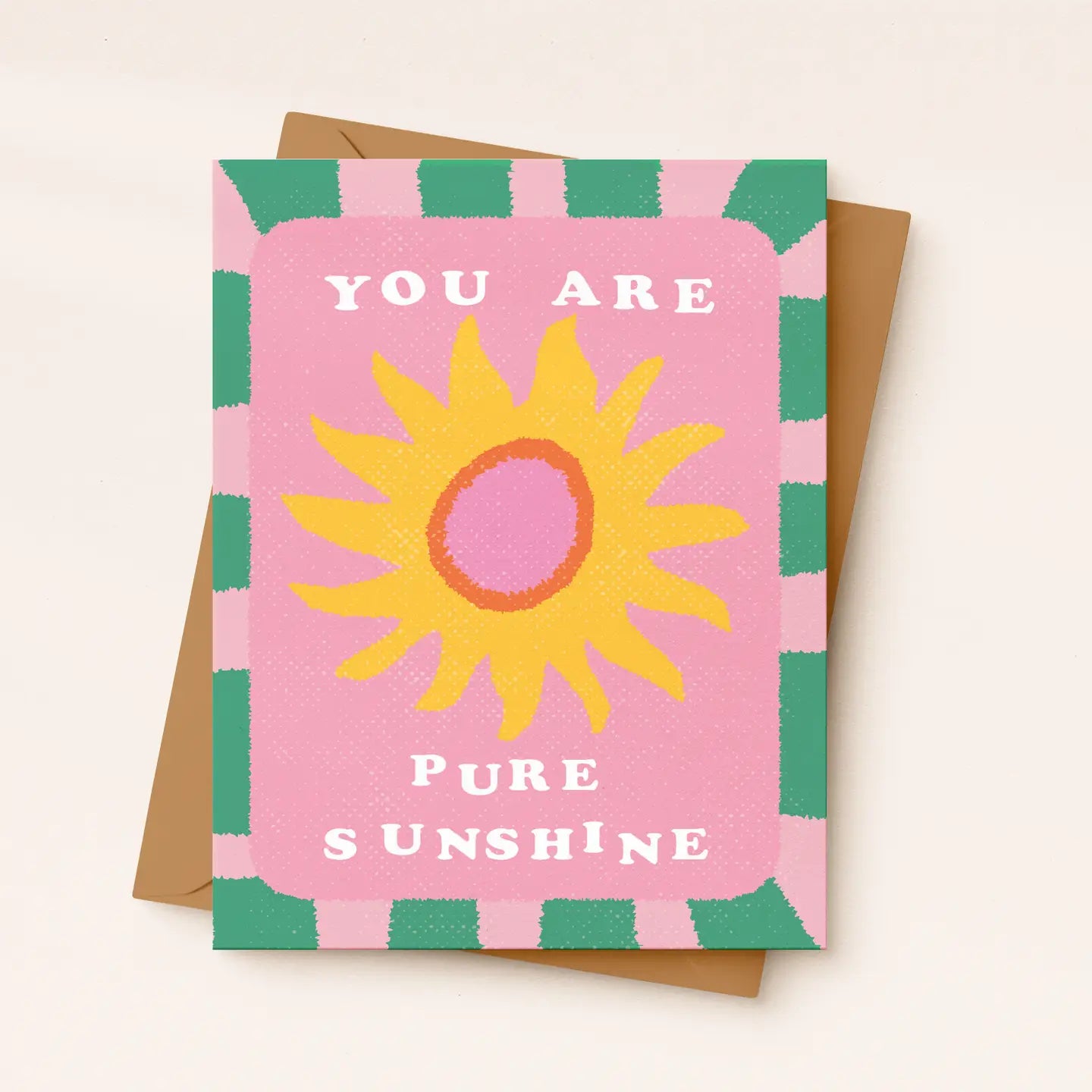 A square greeting card with a pink background, a yellow sun illustration, and the words 'YOU ARE PURE SUNSHINE' in white, shown with a brown envelope on a light beige background.