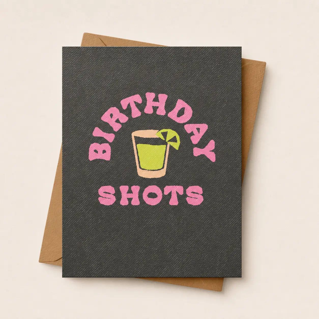 A dark charcoal gray textured greeting card with a pink 'BIRTHDAY SHOTS' graphic and a cocktail illustration, shown with a brown kraft envelope on a light beige background.
