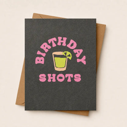 A dark charcoal gray textured greeting card with a pink 'BIRTHDAY SHOTS' graphic and a cocktail illustration, shown with a brown kraft envelope on a light beige background.