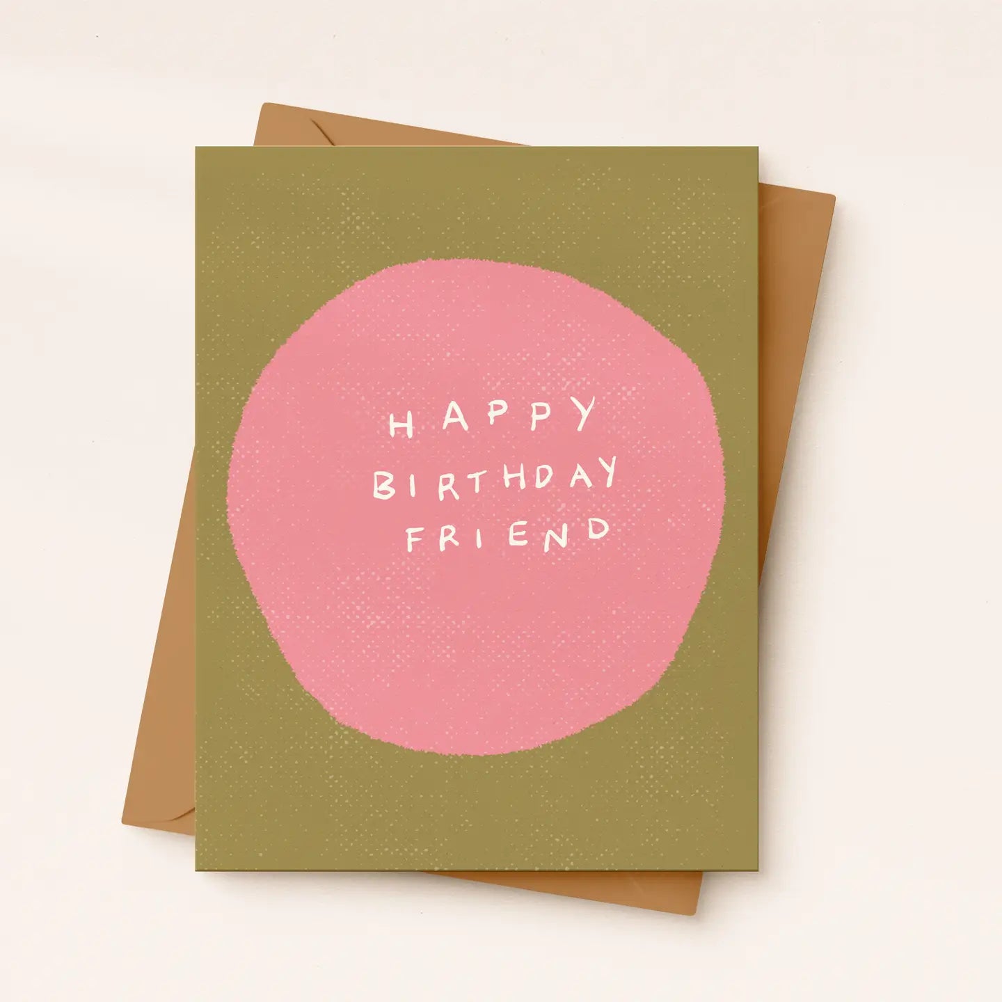 A square greeting card with a muted olive green background, a large pink circle in the center, and white text reading “HAPPY BIRTHDAY FRIEND.” A plain brown envelope is partially visible behind the card on a light warm beige background.