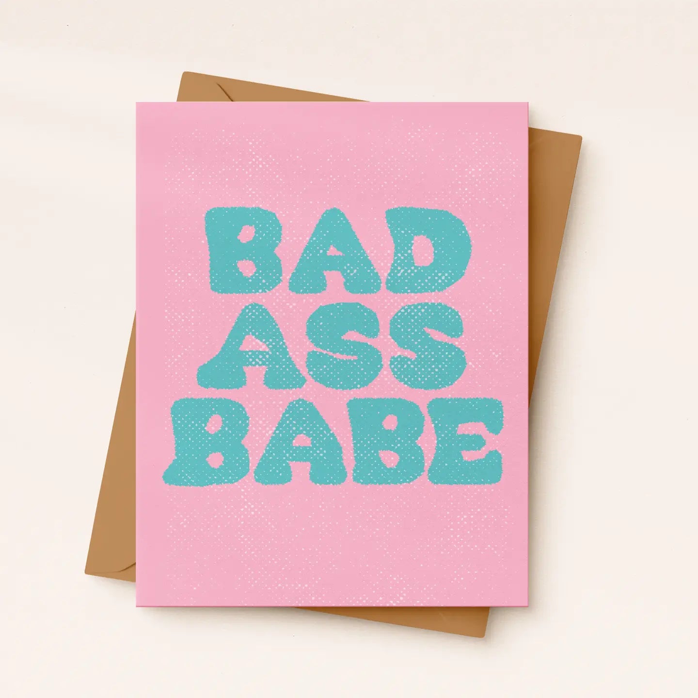 A pink greeting card with the phrase 'BAD ASS BABE' printed in large turquoise letters, shown with a brown envelope on a light beige background.