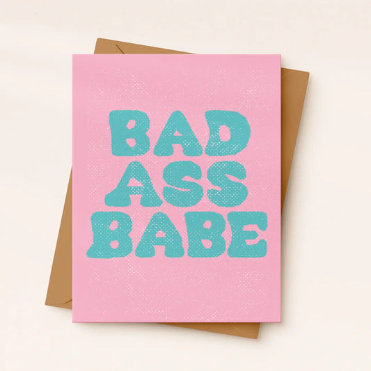 A pink greeting card with the phrase 'BAD ASS BABE' printed in large turquoise letters, shown with a brown envelope on a light beige background.