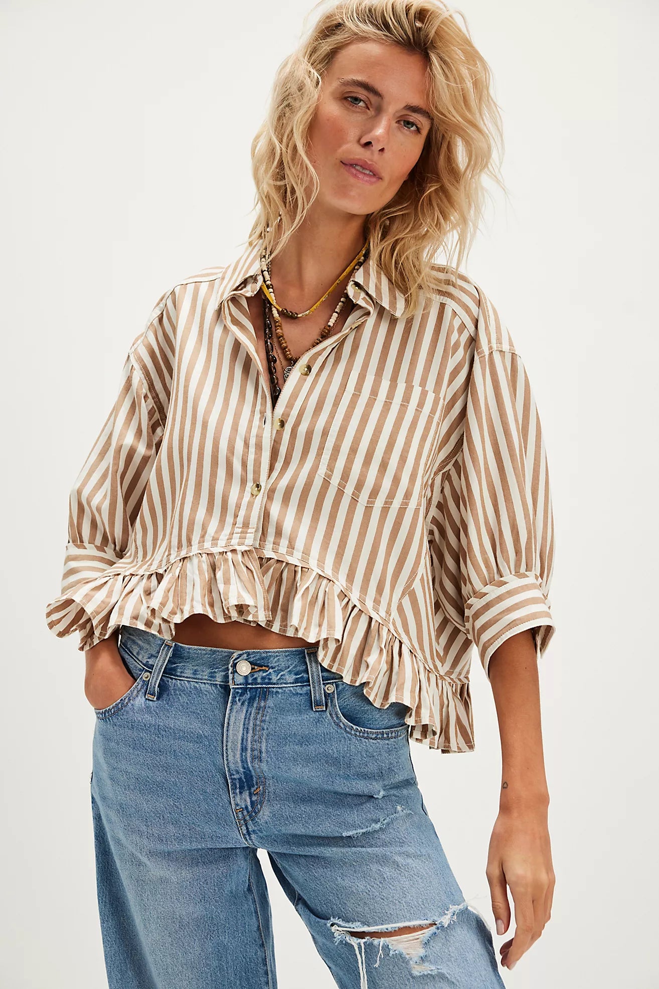 Model wearing a tan and white striped ruffle button-down shirt with light wash jeans and a straw cowboy hat sitting on a wooden chair