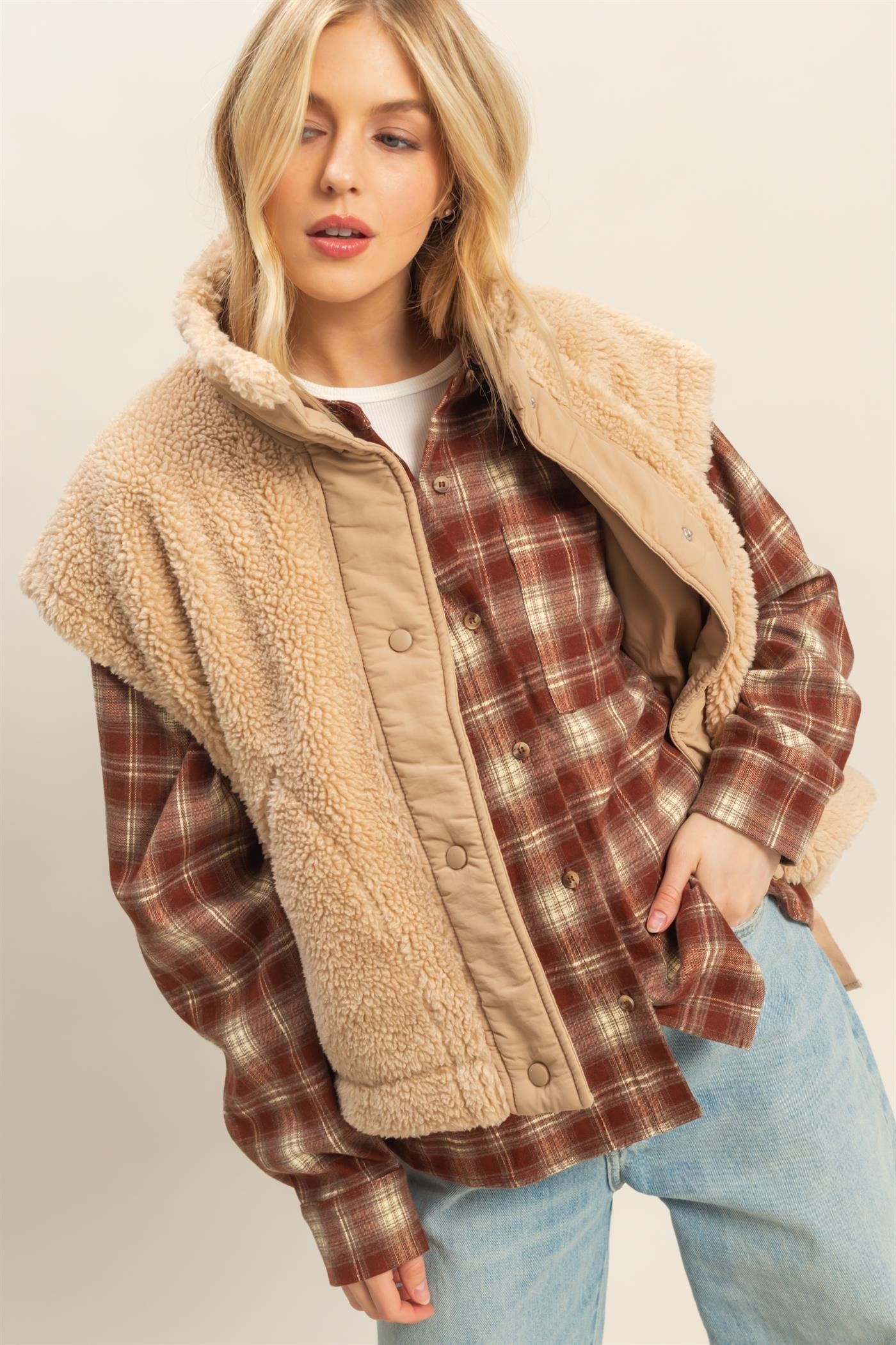 Model wearing beige sherpa vest with snap button placket, tie-up side details, and cap-sleeve effect layered over plaid shirt.