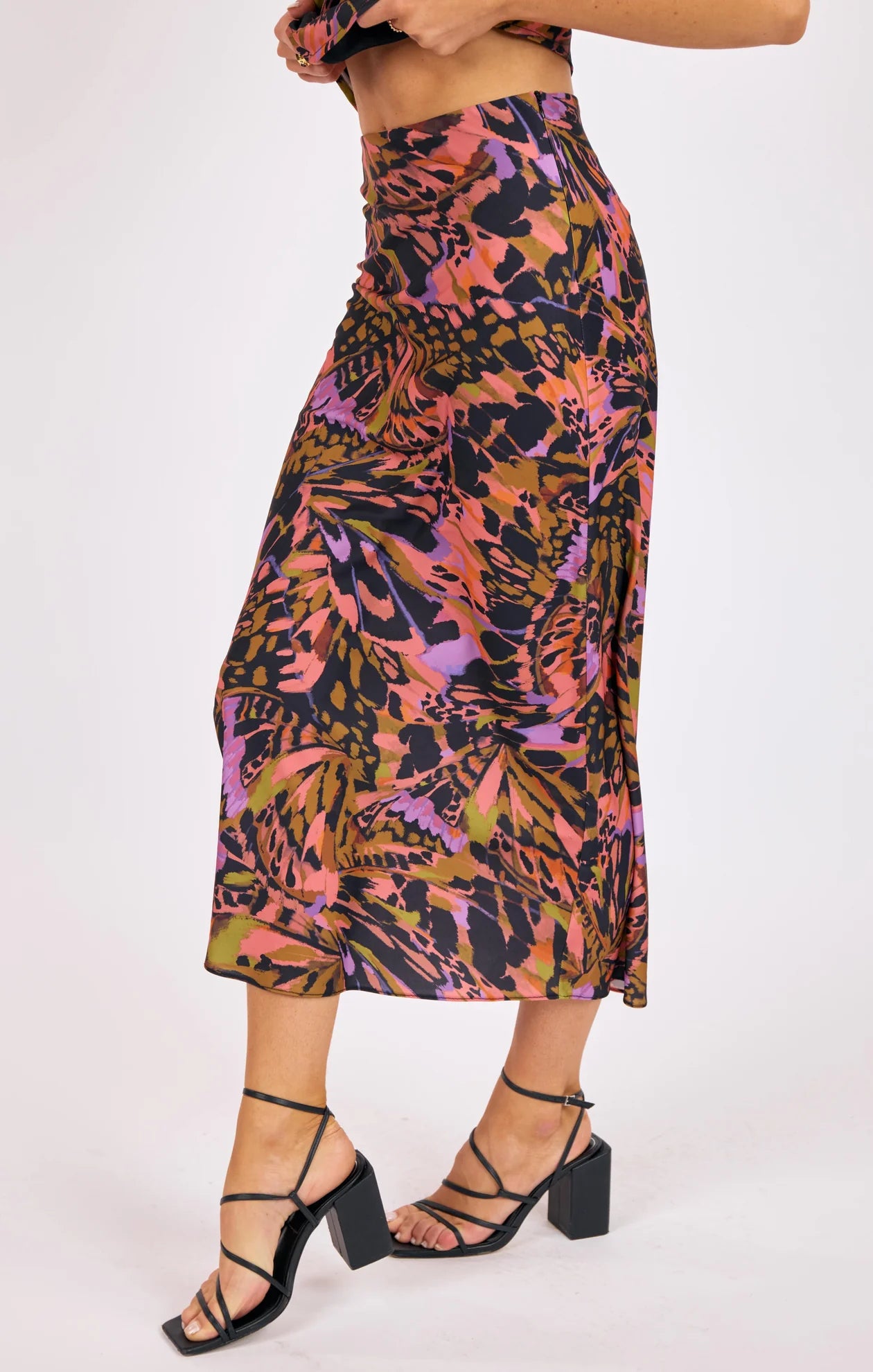 Woman wearing a vibrant butterfly-print satin bias-cut midi skirt paired with strappy black heels.