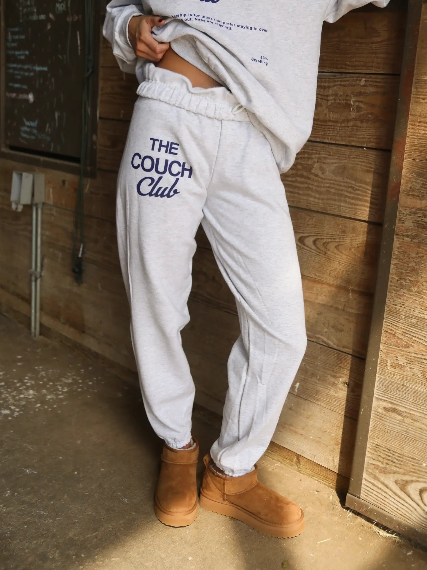 Woman wearing ash grey fleece joggers with “The Couch Club” blue graphic text styled with chestnut slippers.