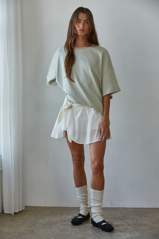 Woman wearing oversized pale green cotton T-shirt with pink contrast neckline, styled with white wrap skirt and ballet flats