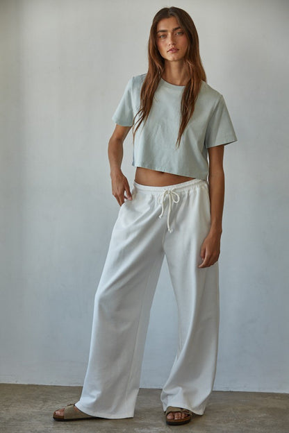 Woman wearing pale blue cropped cotton tee with wide-leg white drawstring pants and slide sandals