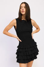 Load image into Gallery viewer, Woman wearing a sleeveless black smocked mini dress with a tiered ruffle skirt, hands on hips, standing against a white background.