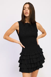 Woman wearing a sleeveless black smocked mini dress with a tiered ruffle skirt, hands on hips, standing against a white background.